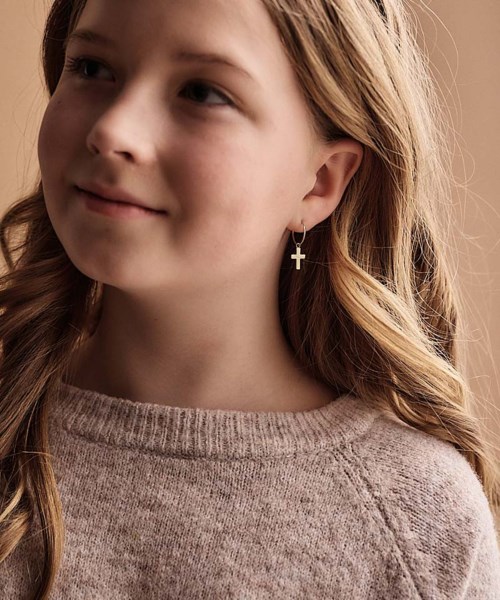 Ung flicka med långt blont hår, bär korsörhängen och en beige tröja, tittar åt sidan med ett mjukt leende.