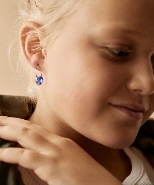 A young girl with blonde hair wearing a blue earring, smiling gently and resting her hand on her shoulder.