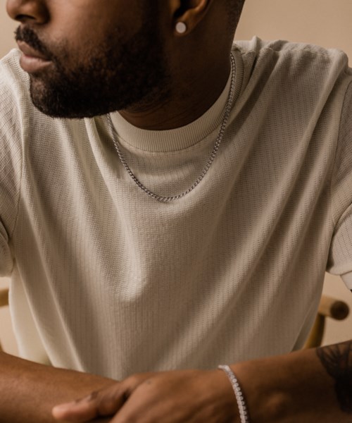 Close-up of a man in a beige shirt with a chain, tattooed arm, and earring, sitting indoors.
