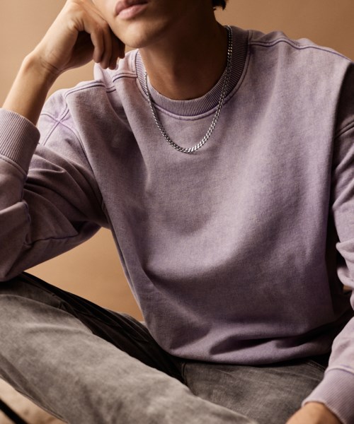 A young man wears a lavender sweatshirt, silver chain necklace, and sits with his hand on his face, against a beige background.