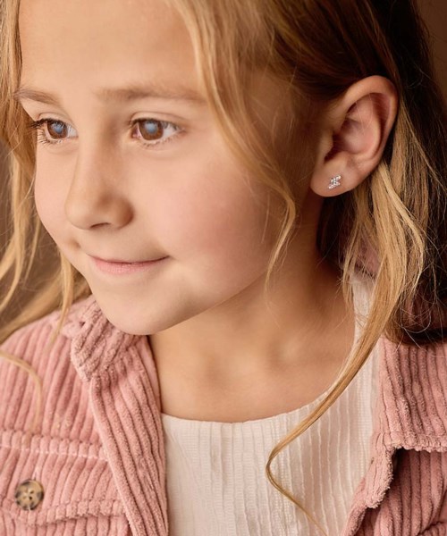 En ung flicka med blont hår, bär en rosa jacka och örhängen, tittar åt sidan.