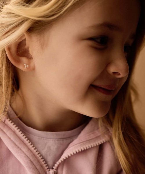 A young girl smiling softly, with blonde hair and small earrings, wearing a light pink zip-up top.