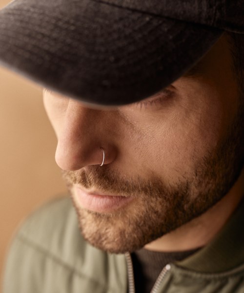 A man with a beard wears a black hat and clothing, with a nose ring, in warm light.