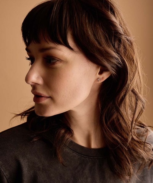 A woman with wavy brown hair and bangs, wearing a dark shirt, looking to the side against a warm beige background.