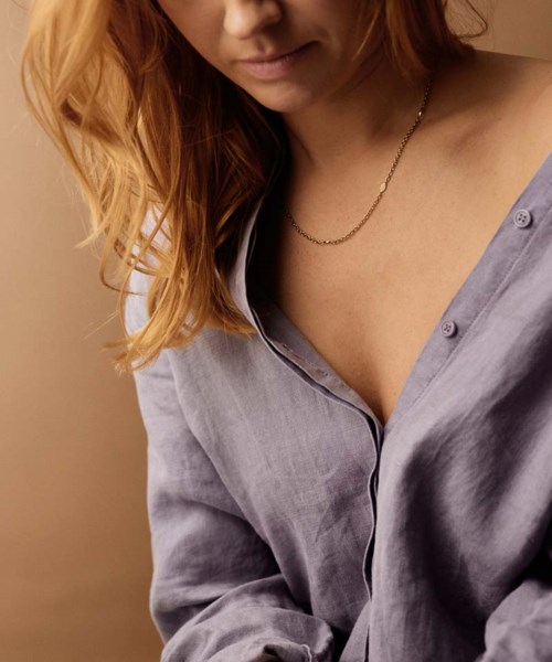 A woman with red hair wearing a loose, open gray shirt and a gold necklace, looking down slightly.