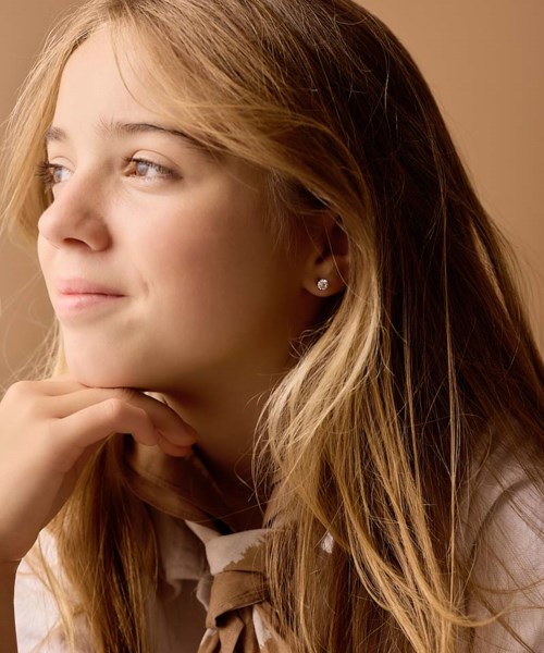 A young girl with long blonde hair, looking thoughtfully to the side, smiling slightly, wearing small earrings.