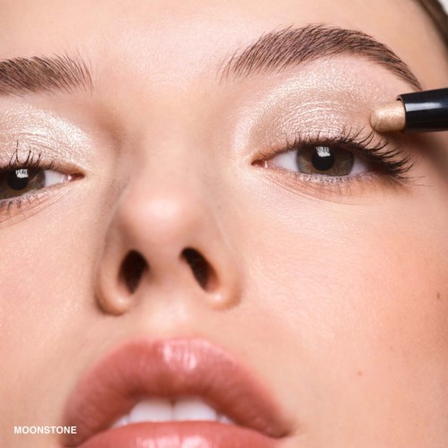 A woman smudges gold makeup on her eyelid with a brush, close-up of her face showing detailed eye makeup.