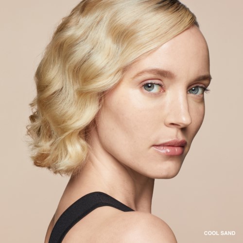 A woman with wavy blonde hair and blue eyes looks over her shoulder, wearing a black top and neutral makeup.