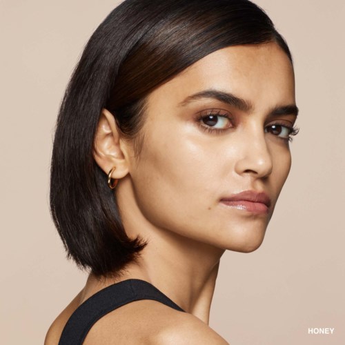 A woman with straight dark hair and gold earrings, looking slightly to the side, against a beige background.
