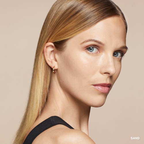 A woman with long blonde hair, fair skin, blue eyes, small earrings, and natural makeup looks over her shoulder.