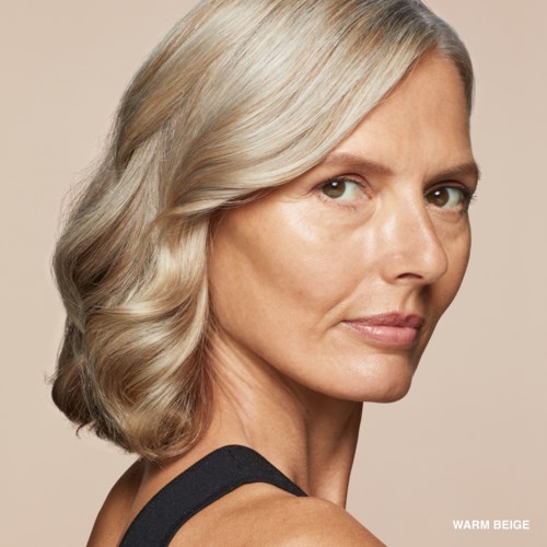 A woman with blonde, wavy hair looks over her shoulder on a beige background.