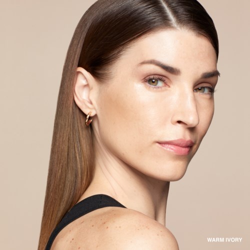 A woman with straight brown hair, fair skin, wearing small earrings and a black top, looking over her shoulder.