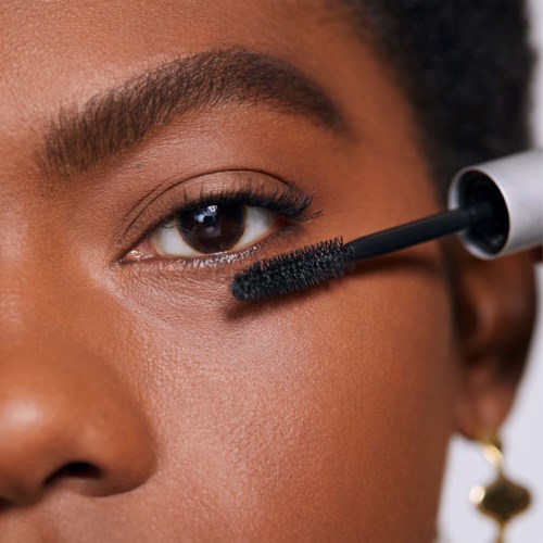 A woman applies mascara to her eye, focusing on her eyebrow and nearby skin. She has smooth, brown skin and wears a small earring.