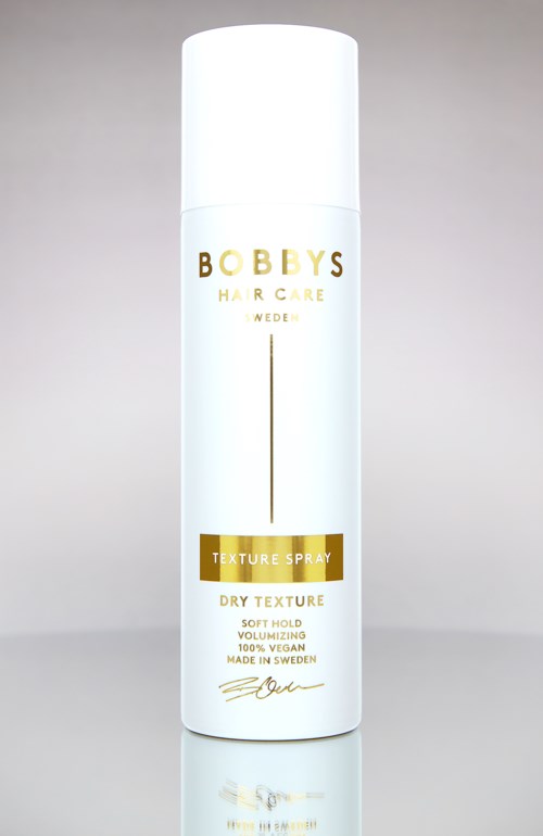 Witte cilindervormige fles met goud en zwart tekst, labelt als textuurspray voor droog haar. Vermeldt ook veganistisch en gemaakt in Zweden.