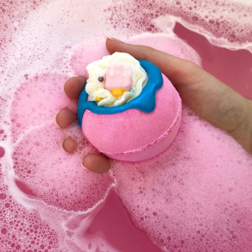 En person håller en rosa badbomb med en kycklingansikte över rosa bubbligt vatten.