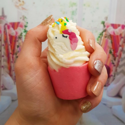 Een hand houdt een roze cupcake met witte glazuur, een eenhoorn-decoratie en kleurrijke sprinkles.