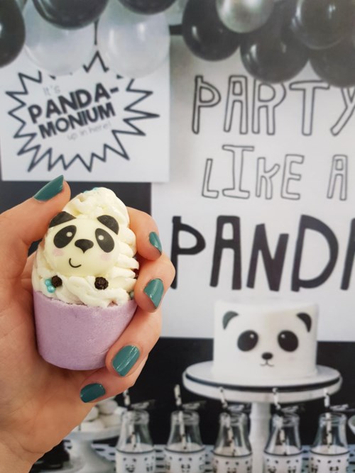 Een panda-thema taart in een paarse omhulsel, vasthouden door een hand, met panda-decoraties en ballonnen op de achtergrond.