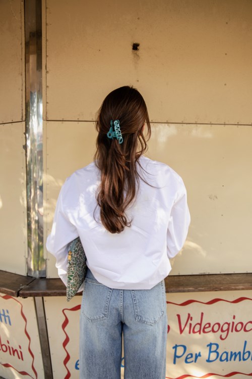 A girl with long hair, a blue hair clip, white jacket, and light jeans stands facing away near a wall.