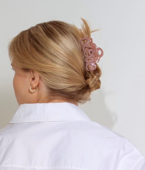 Woman with blonde hair styled in an updo, secured with a pink hair accessory, wearing earrings and a white shirt.