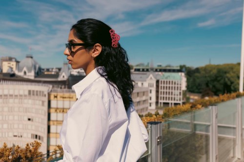 A woman with dark hair, sunglasses, and a pink hair accessory stands outdoors on a sunny day, overlooking city buildings.