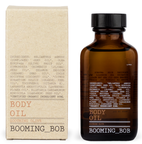 Eine braune Flasche mit der Aufschrift 'Body Oil' und einem orangefarbenen 'Soothing Olive'-Etikett steht neben einer beigefarbenen Verpackung mit Zutatenliste.