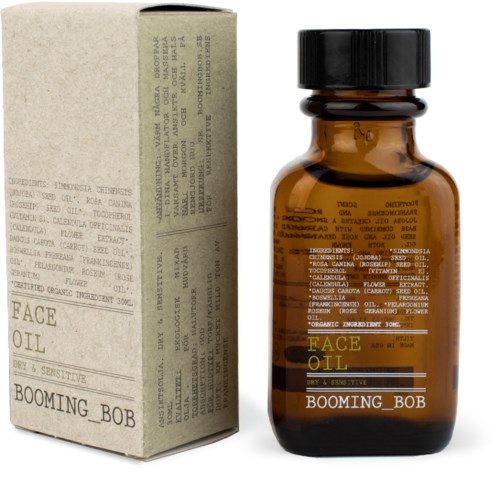 Kleine braune Glasflasche mit Beschriftung 'FACE OIL' und 'BOOMING_BOB' neben einem beigen Karton mit Text und Anweisungen.