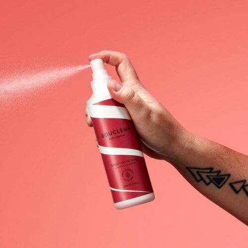 Een hand spuit een roze fles met witte en roze strepen tegen een roze achtergrond.