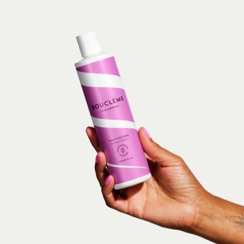 Een hand houdt een roze en witte fles met het label Bouclème, die voor haarverzorging is.