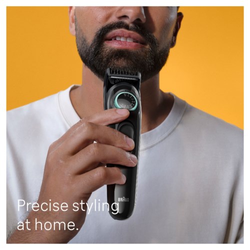 A man with a beard uses an electric shaver, standing against a yellow background, wearing a white shirt.