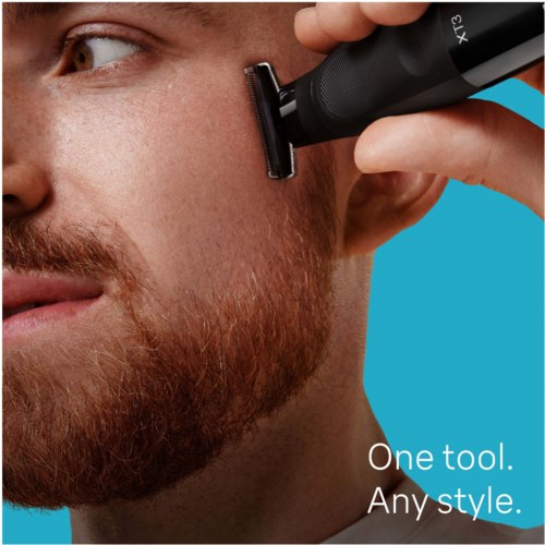A man shaves his face with a black electric razor. He has a beard and looks to the side. Blue background.