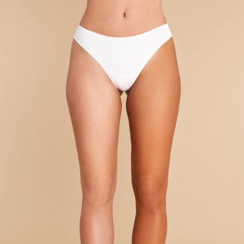 Een vrouw draagt witte ondergoed, staat tegen een beige achtergrond, met focus op haar onderlichaam.
