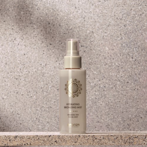 A white spray bottle labeled 'Hydrating Bronzing Mist' stands on a textured surface, with a plain gray background.