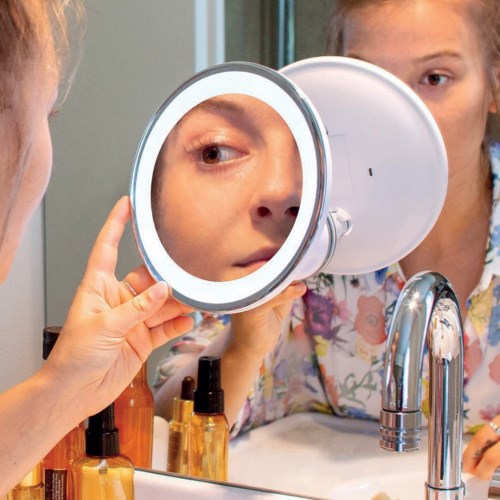A girl peering into a round mirror while another girl looks on, with bottles and a sink visible nearby.