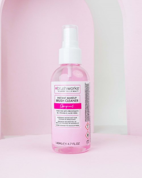 A pink bottle of makeup brush cleaner with a spray top, placed against pink and white background.