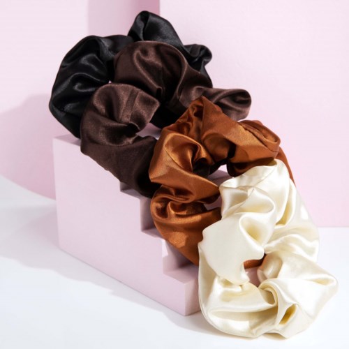 En uppsättning av fyra satin-scrunchies i svart, brunt, rostfärgat och krämfärgat, arrangerade på en rosa kub mot en rosa bakgrund.