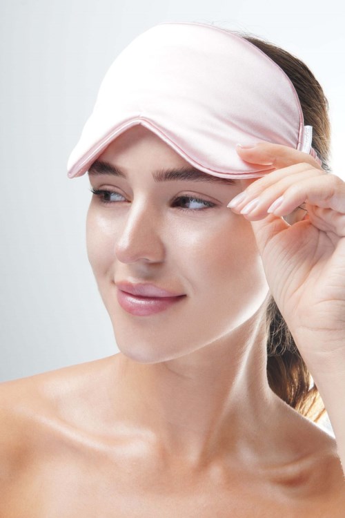 A woman adjusts a pink sleep mask on her forehead, smiling softly, with clear skin and brown hair.