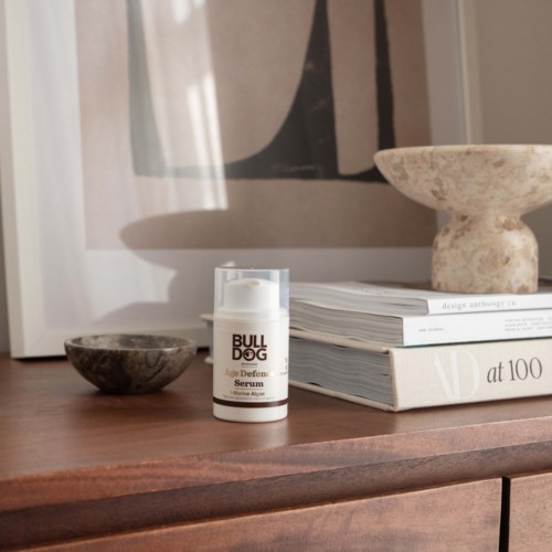 Eine Flasche Bulldog Age Defence Serum auf einer Holzoberfläche mit Büchern und Dekorationssteinen.