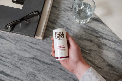 Käsi pitää deodoranttia merkinnällä 'BULLDOG' pöydällä, jossa on laseja ja kirjoja.