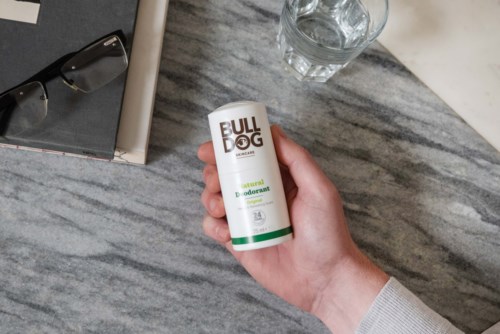 A hand holds a BULLDOG natural deodorant stick near a glass of water and eyeglasses on a gray marble surface.