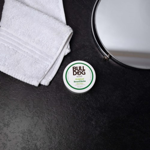 A white towel, a round mirror, and a container of beard balm on a dark surface.