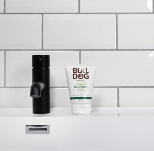 Een zwarte zeepdispenser en een tube Bulldog Original moisturizer op een wit wastafelkastje met witte tegels achter.