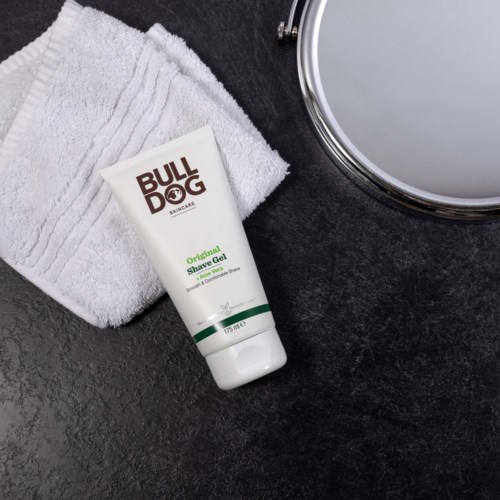 A white tube of Bull Dog shave gel lies on a dark surface next to a folded white towel and a round mirror.