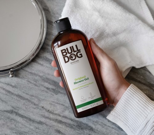 Eine Hand hält eine braune Flasche Bulldog Skincare Duschgel, neben einem Spiegel auf Marmor.