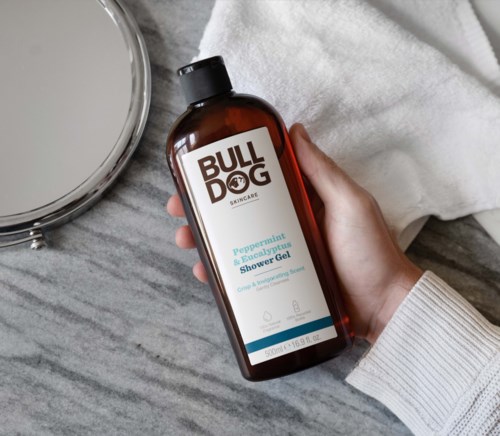 Eine Hand hält eine braune Flasche mit "BULLDOG" Aufschrift und Duschgel, neben einem Spiegel und einem weißen Handtuch.