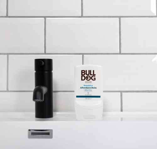 Een zwart zeeppompje en een witte tube met 'Bull Dog' op een witte plank tegen een betegelde muur.