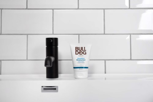 Een zwarte zeepdispenser en een witte tube met "BULLDOG" staan op een wit wasbakje met betegelde muur erachter.