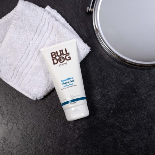 A tube of Bulldog Sensitive Shave Gel placed near a white towel and a mirror on a dark surface.