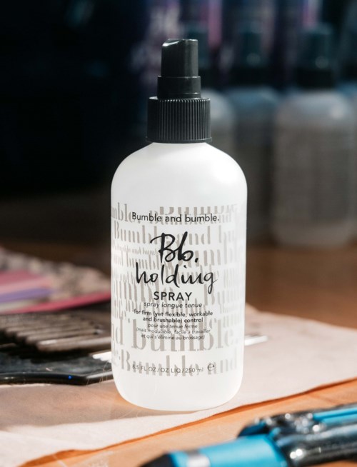 Der Sprayflasche mit der Aufschrift 'Bumble and bumble. Blow holding Spray' steht auf einem Tisch mit anderen Gegenständen im Hintergrund.