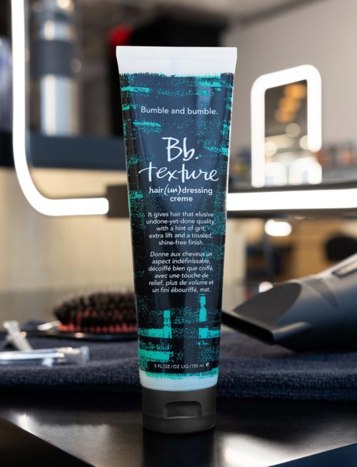 Eine Tube Bumble and bumble Haarkrem auf einer dunklen Oberfläche mit einem Spiegel und einer Haarbürste im Hintergrund.
