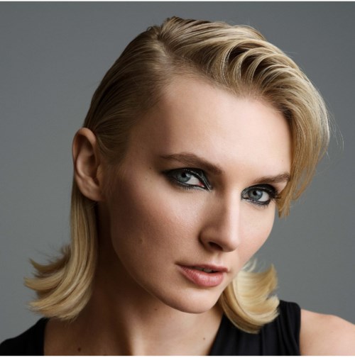 Eine Frau mit blondem Haar und blauen Augen schaut selbstbewusst in die Kamera.